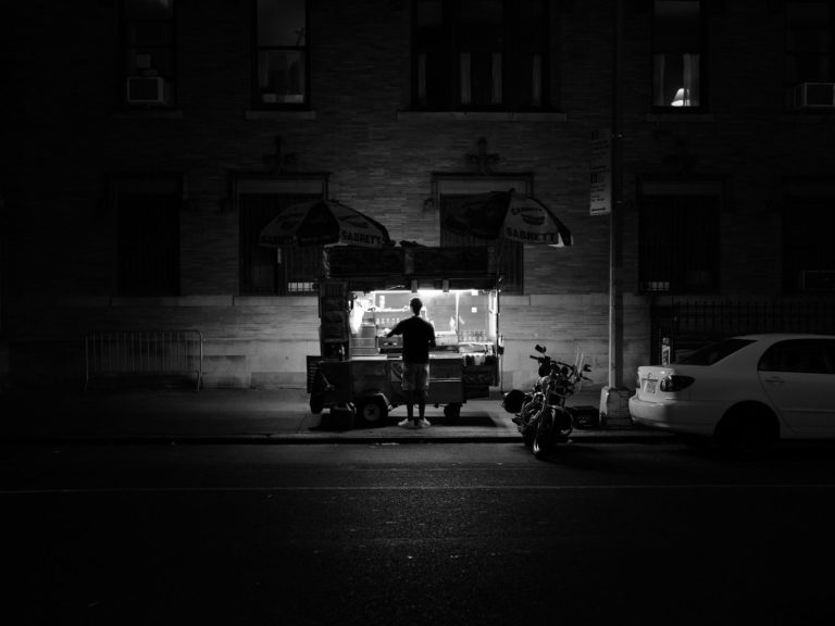 Ein Straßenverkäufer steht vor einem beleuchteten Imbissstand in der Dunkelheit.