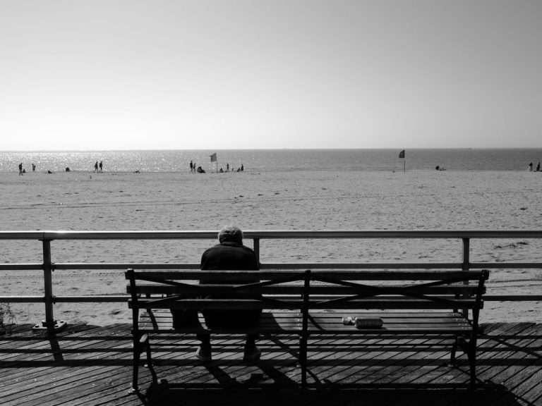 Eine Person sitzt auf einer Bank am Strand und blickt nachdenklich aufs Wasser.